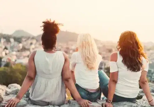 Three women sitting together on a wall, looking out towards sunrise—symbolising resilience, hope, and the importance of supportive relationships in suicide prevention and mental health.