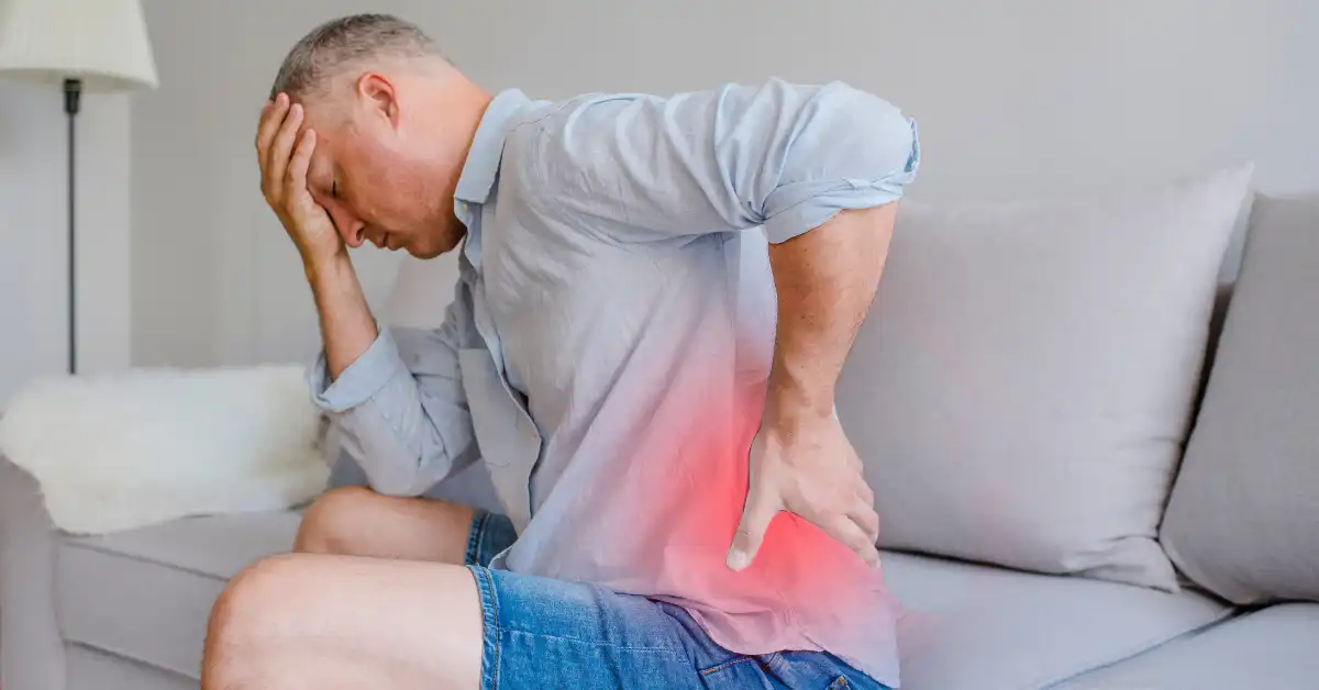 persistent-pain-featured-1200x628 Person sitting on a couch holding their lower back, illustrating the physical and emotional impact of persistent pain