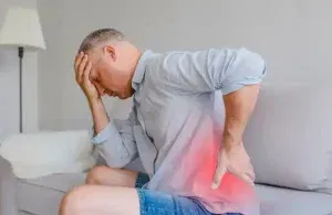Person sitting on couch experiencing persistent pain, highlighting the need for psychological support and pain management