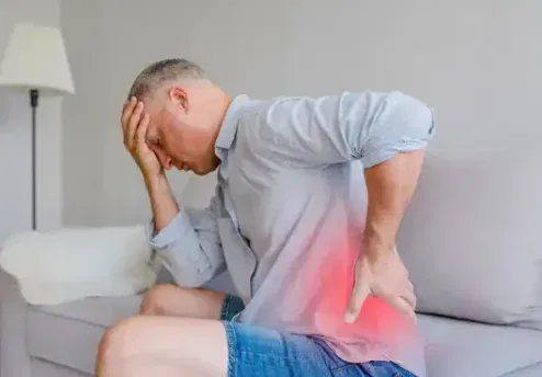 Person sitting on couch experiencing persistent pain, highlighting the need for psychological support and pain management