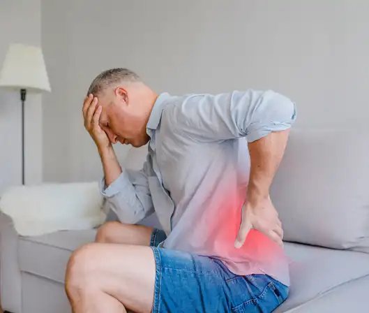 Person sitting on couch experiencing persistent pain, highlighting the need for psychological support and pain management